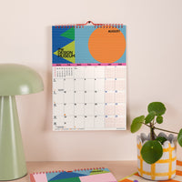 A colorful spiral-bound wall calendar hanging on a beige wall in a styled room setting, featuring a retro design with geometric shapes and the text 'THE DESIGN MUSEUM' and 'AUGUST'. A mint green table lamp is on the left, and a potted green plant in a white and yellow patterned pot is on the right.