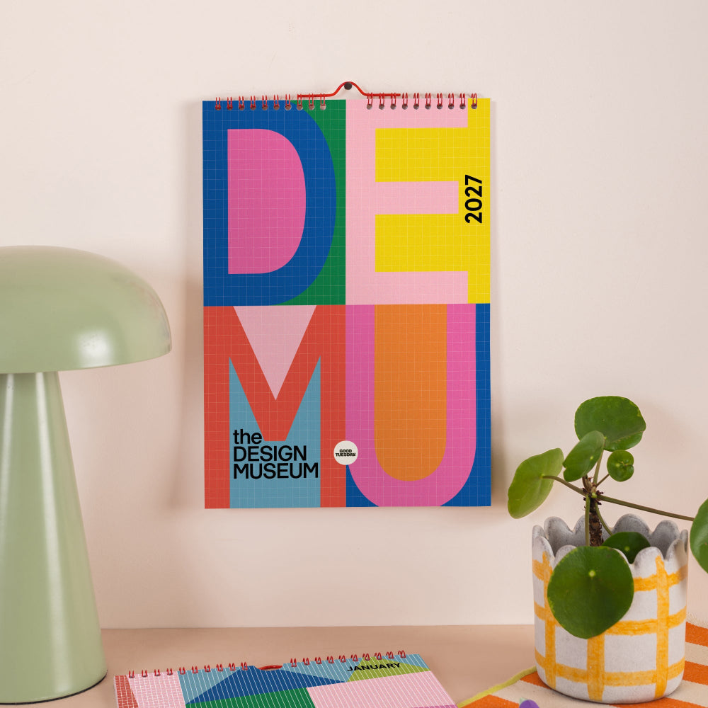 A colorful 2027 wall calendar titled 'DEMU the DESIGN MUSEUM' hanging on a beige wall in a styled room setting with a green lamp, a potted plant, and other notebooks.