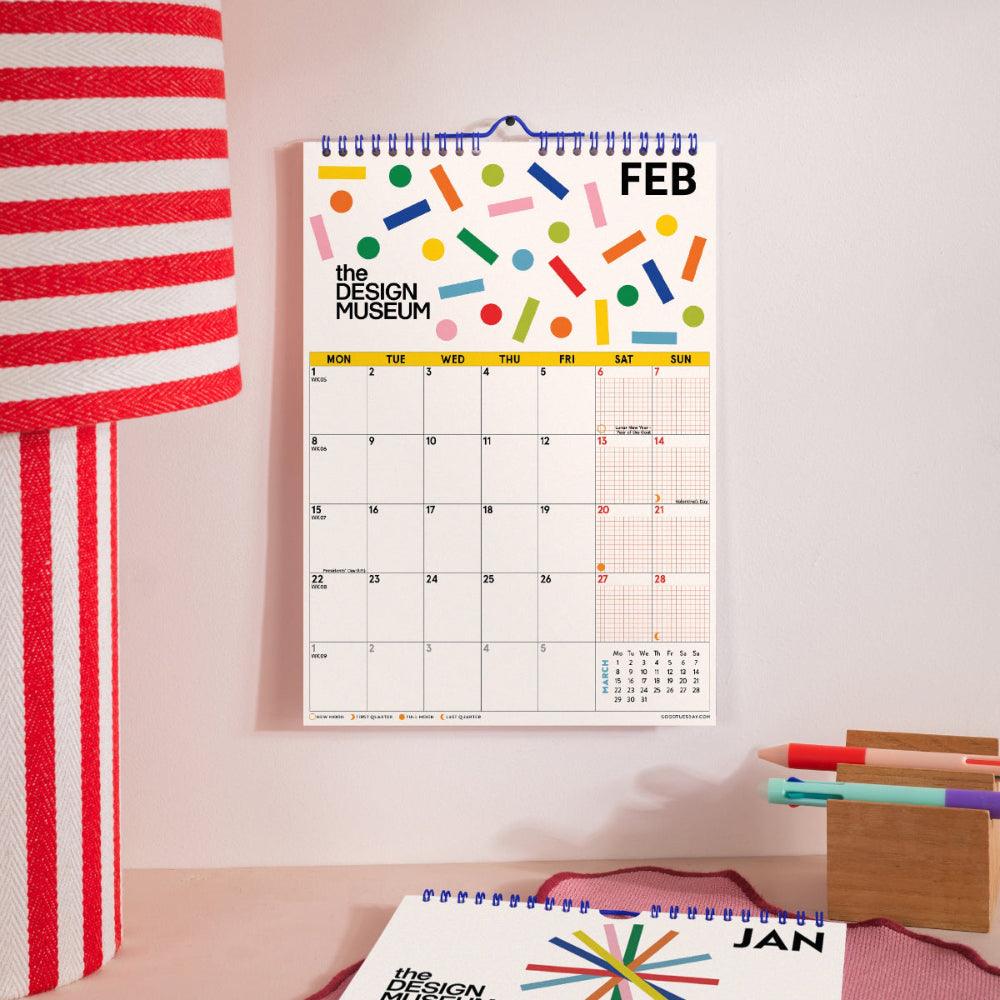A spiral-bound February desk calendar from The Design Museum hangs on a light pink wall. The calendar features a colorful abstract geometric pattern at the top and a clean monthly grid below.