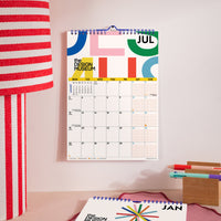 A spiral-bound wall calendar hanging on a light pink wall, showing July with colorful abstract graphics and the text 'the DESIGN MUSEUM.' A matching calendar page for January lies on a desk in the foreground, with a striped lamp on the left and stationery on the right.
