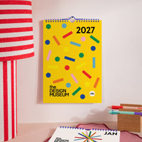A bright yellow 2027 wall calendar hanging on a light pink wall, decorated with colorful confetti shapes. The calendar features the text 'the DESIGN MUSEUM' and '2027'.