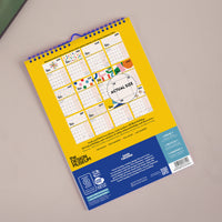 A spiral-bound desk calendar shown from the back, featuring a bright yellow top section with a 12-month grid and colorful decorative illustrations, plus a dark blue bottom section with branding and product details. The calendar is photographed on a neutral beige surface.