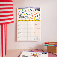 A spiral-bound February desk calendar from The Design Museum hangs on a light pink wall. The calendar features a colorful abstract geometric pattern at the top and a clean monthly grid below.