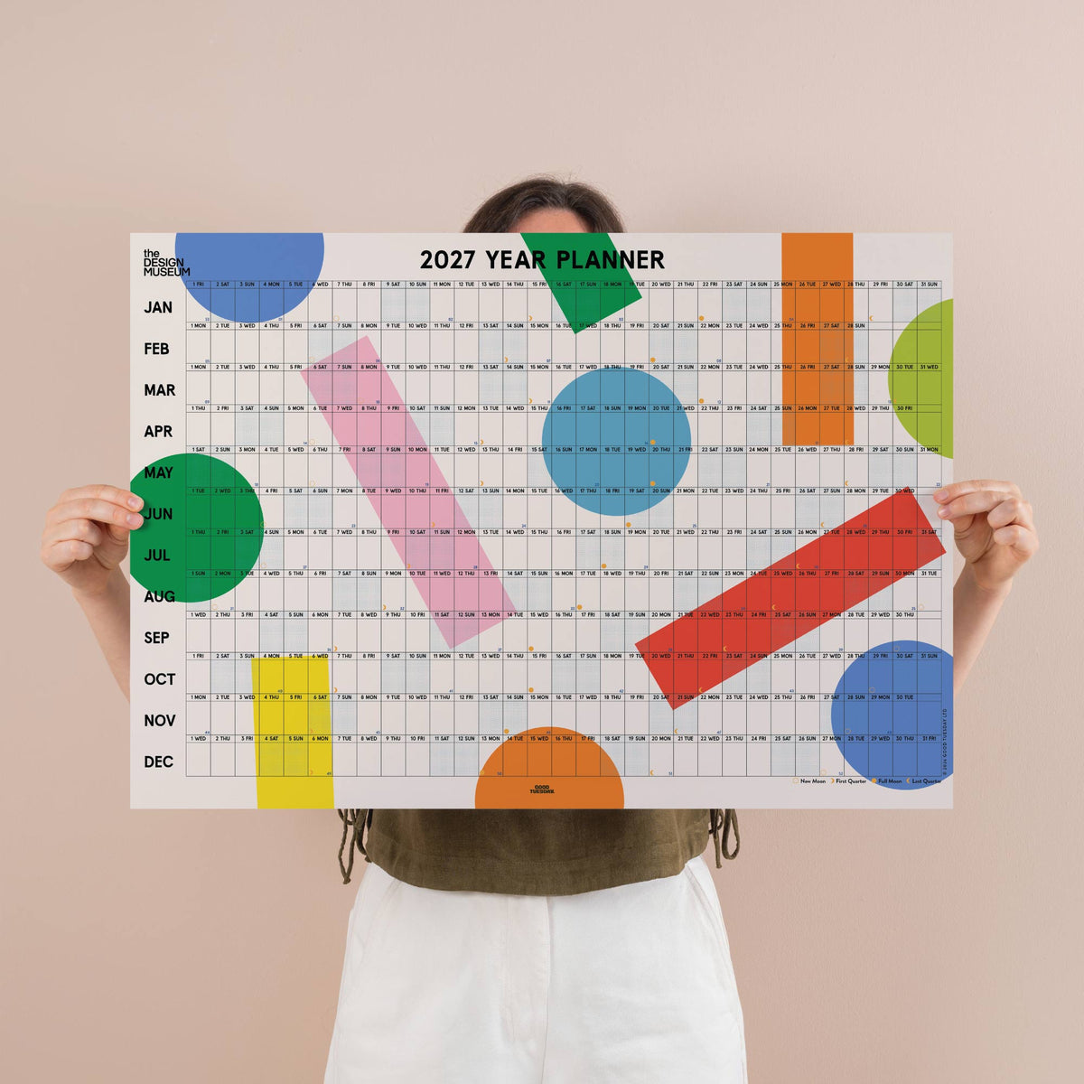 A person holds up a large 2027 Year Planner calendar page against a solid beige background. The planner features a white grid with colorful geometric shapes in blue, green, pink, orange, and red.