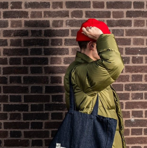 Person wearing a green jacket and red cap, holding a navy tote bag against a brick wall.