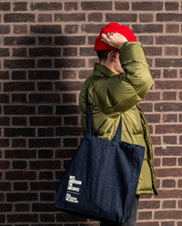 Person wearing a green jacket and red cap, holding a navy tote bag against a brick wall.