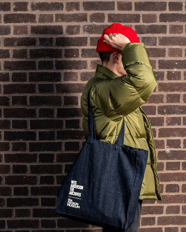 Person wearing a green jacket and red cap, holding a navy tote bag against a brick wall.