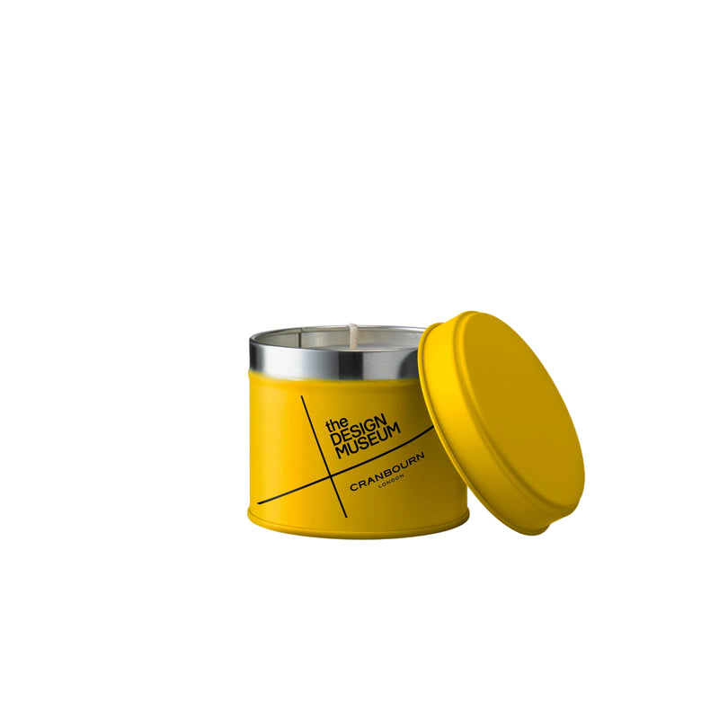 A yellow cylindrical candle tin with its lid leaning against it, featuring black text that reads 'THE DESIGN MUSEUM' and 'CRANBOURN DESIGN'. The tin has a silver metal rim at the top and is photographed on a clean white background.