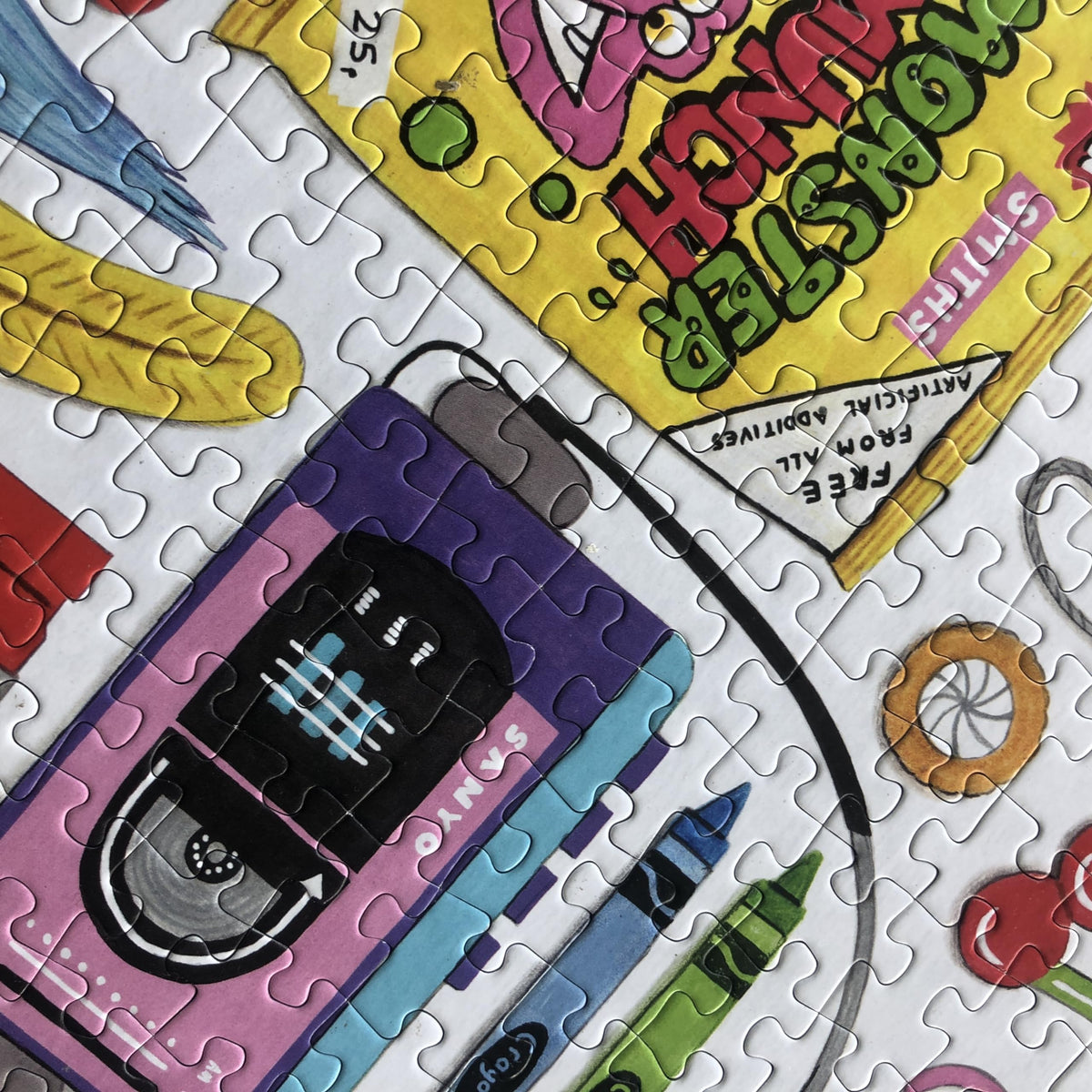A closeup view of a 1990s themed jigsaw puzzle featuring colourful illustrations of a cassette tape and crayons.