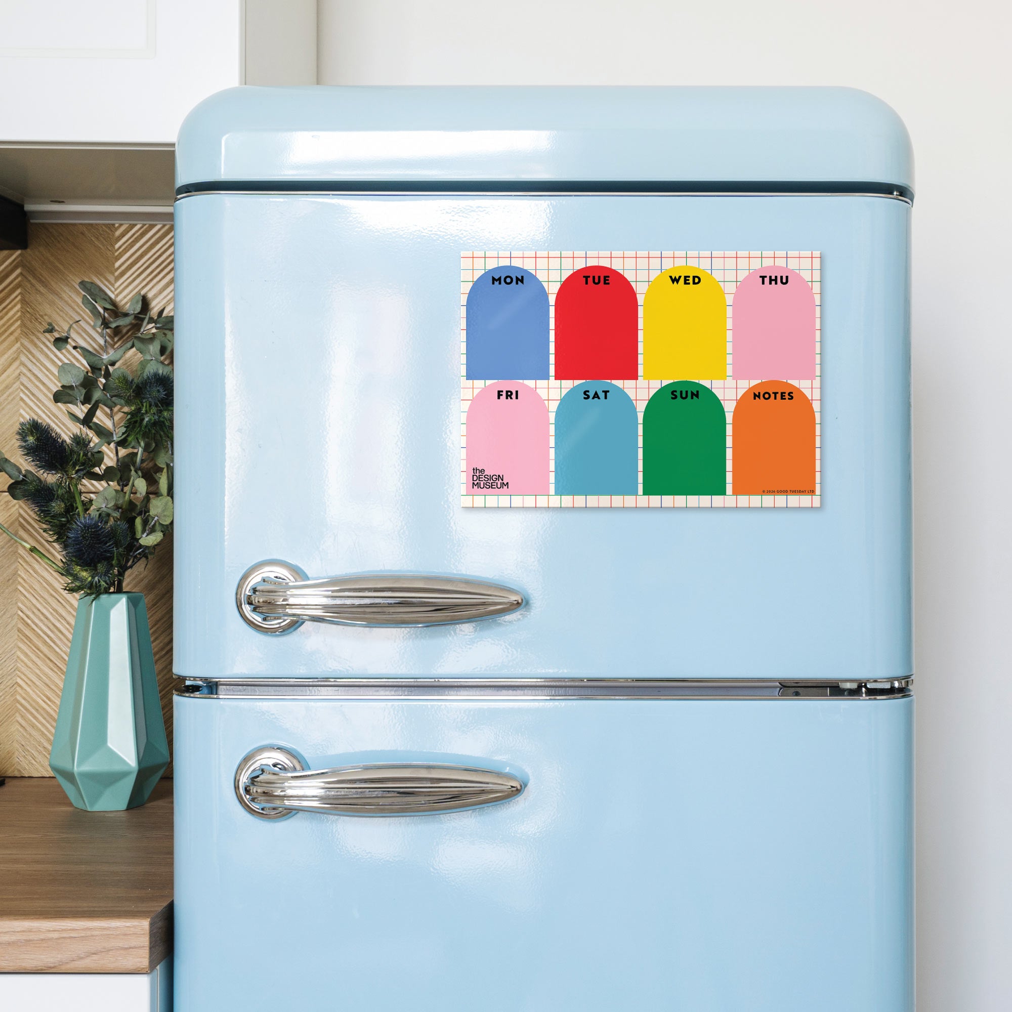 A pastel blue retro-style refrigerator in a kitchen setting, featuring a colorful weekly planner magnet on the upper door. The magnet displays days of the week in arched color blocks with labels 'MON', 'TUE', 'WED', 'THU', 'FRI', 'SAT', 'SUN', and 'HOLY'.