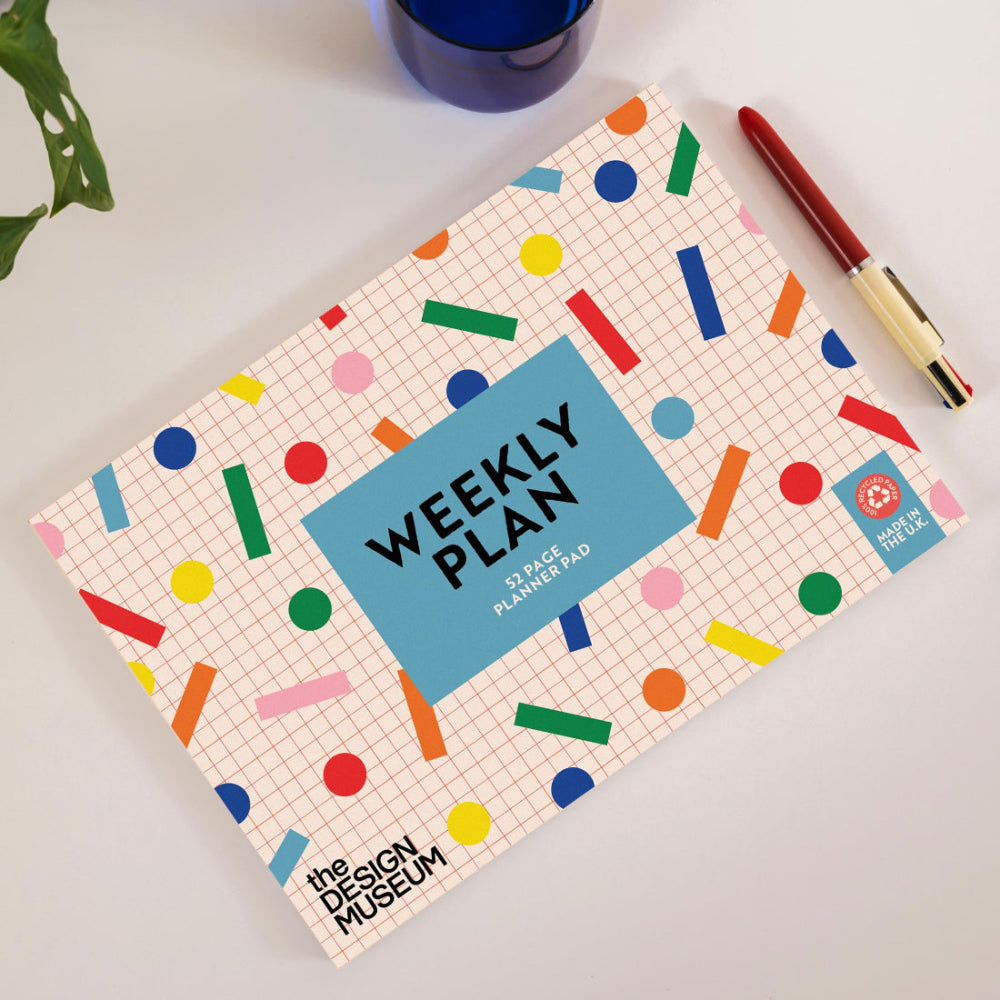 A weekly planner cover laid flat on a light surface, featuring a pastel pink grid background with colorful confetti dots and bars, a large blue center label reading 'WEEKLY PLAN,' and a small 'the DESIGN MUSEUM' logo. A red pen rests beside it, with a blue cup and green plant leaves partially visible around the scene.