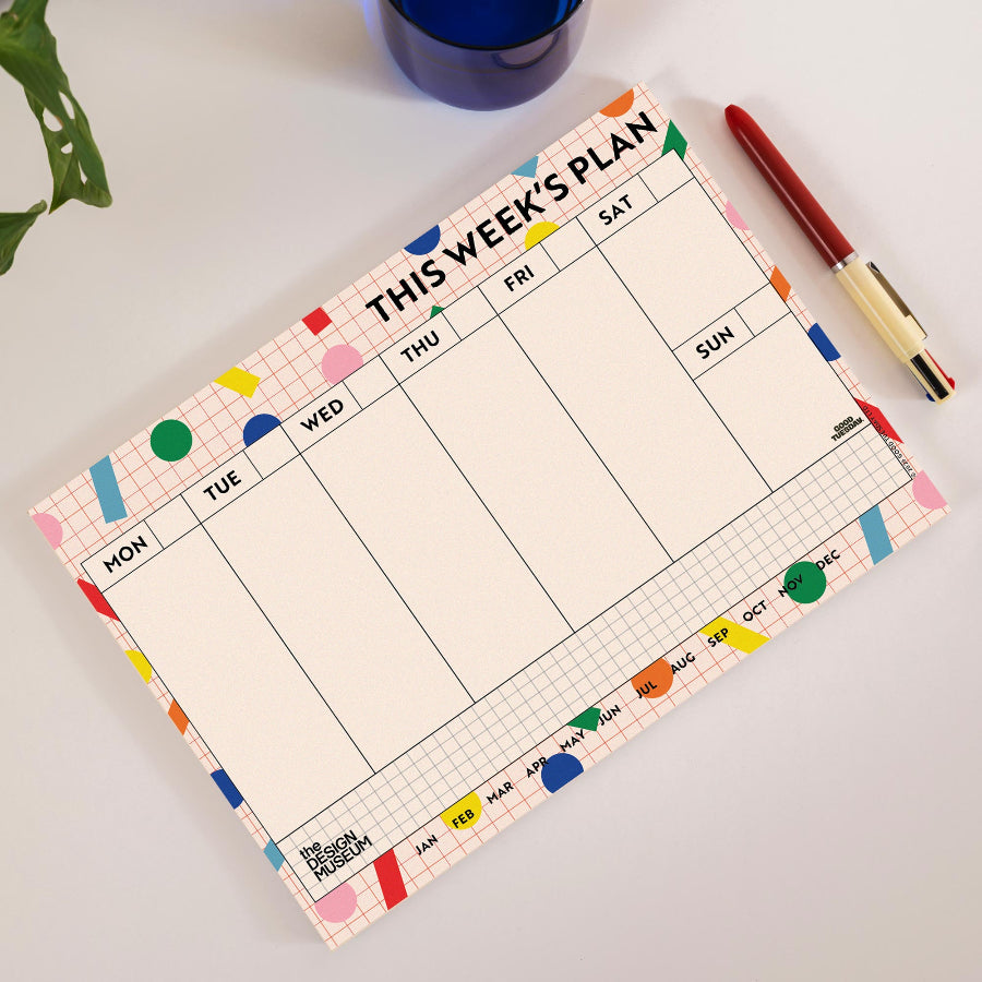 A weekly planner pad with a colorful grid layout and decorative pastel accents, shown on a light surface next to a red pen and a blue glass.