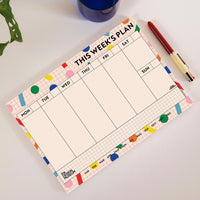 A weekly planner pad with a colorful grid layout and decorative pastel accents, shown on a light surface next to a red pen and a blue glass.