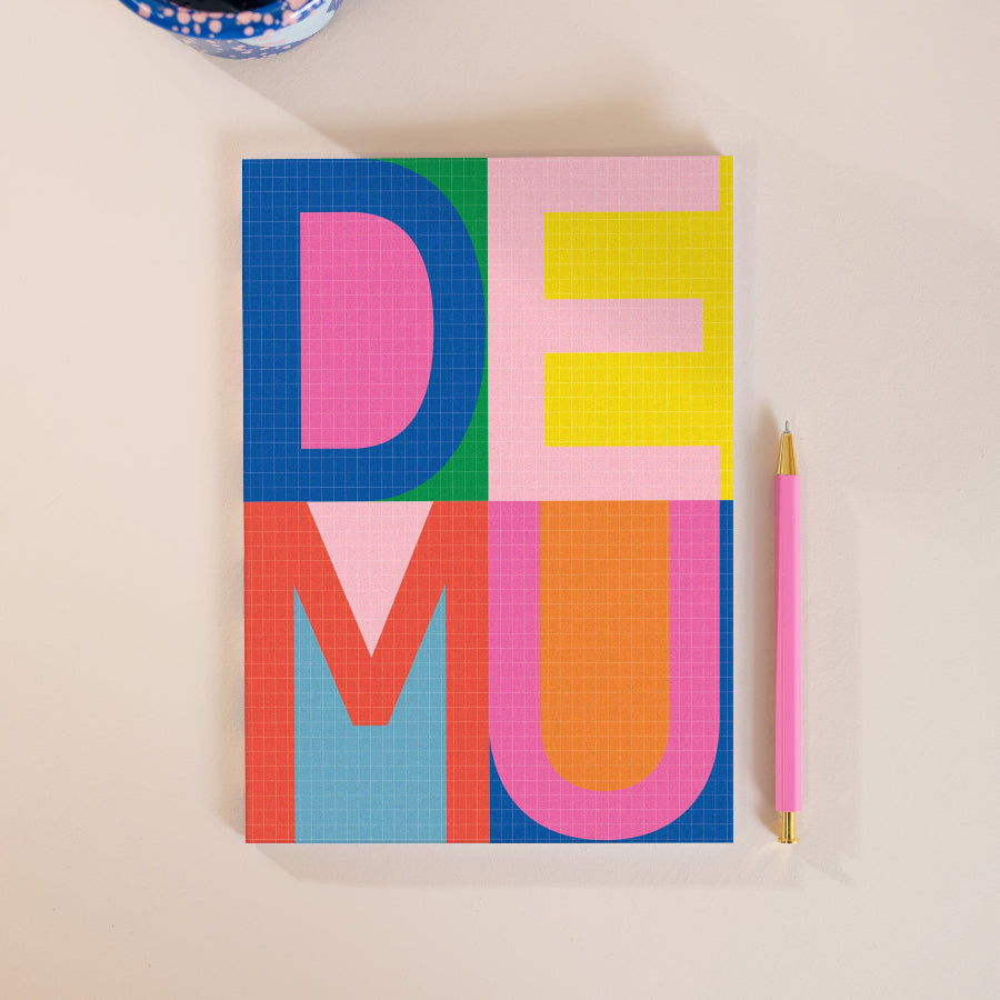 A colorful typographic art print featuring the word 'DEMU' in large block letters on a grid pattern, styled on a light beige surface next to a pink pen.