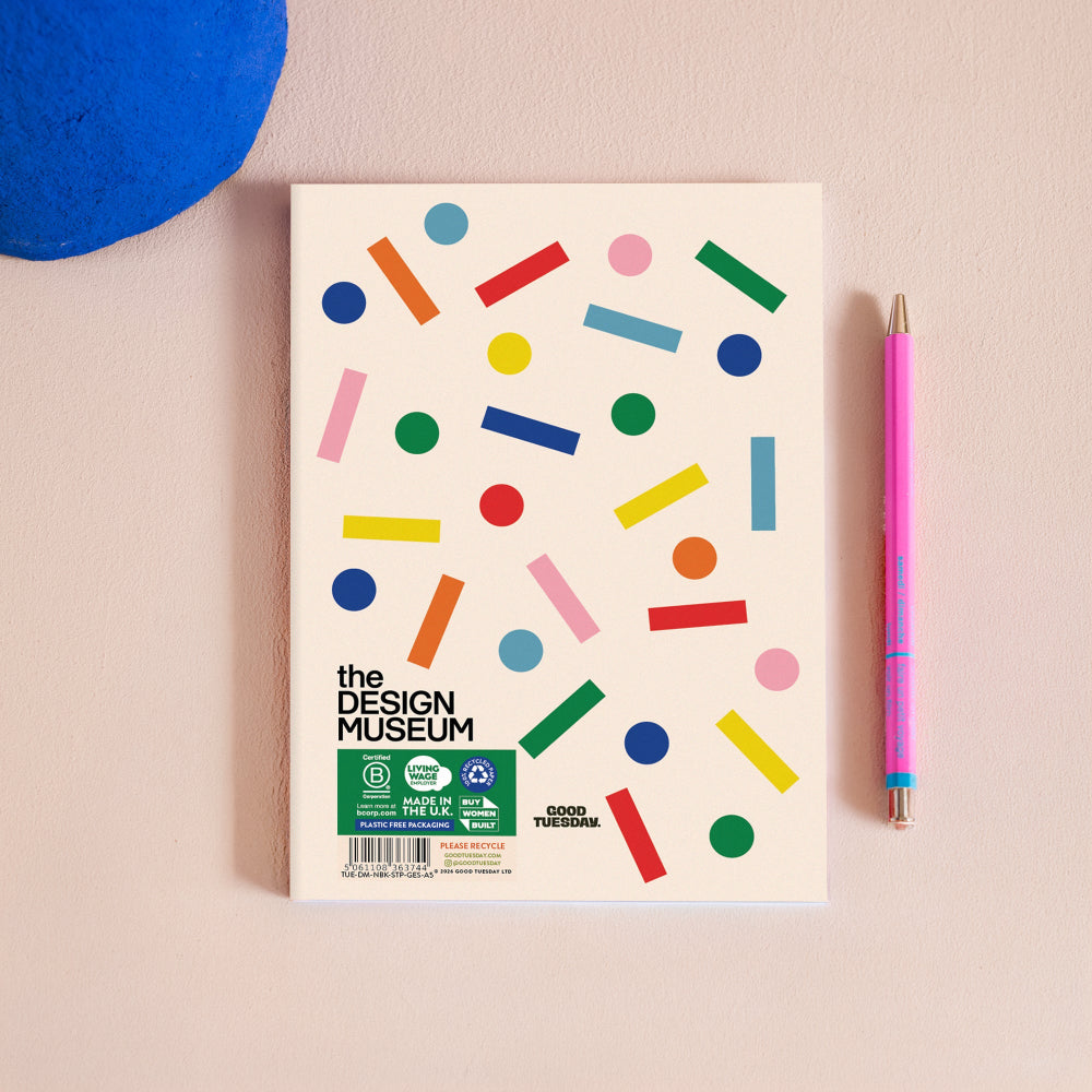 A notebook with a cream-colored cover decorated with colorful geometric shapes, placed on a light beige surface next to a pink mechanical pencil. The cover features the text 'the DESIGN MUSEUM' and a green 'GOOD TOUCHES' logo at the bottom left.