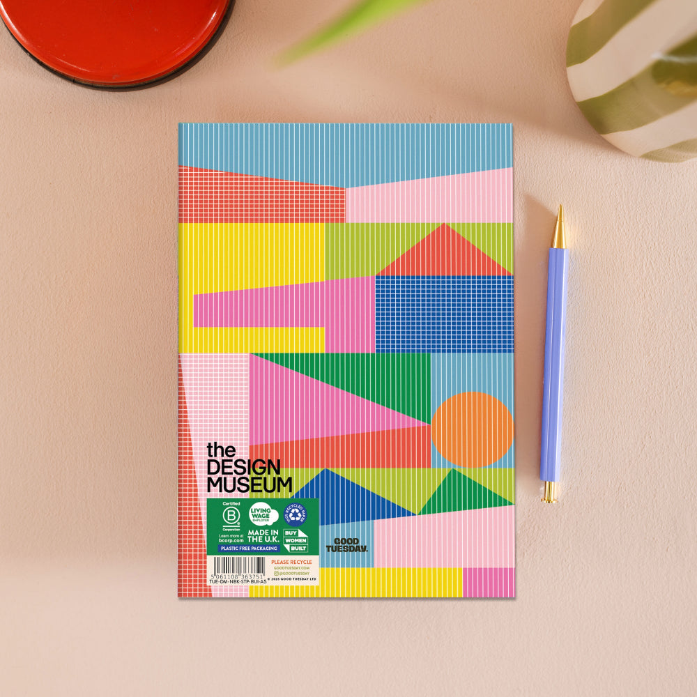 A colorful notebook on a beige surface next to a blue pen. The notebook cover features a vibrant geometric pattern with pink, yellow, green, blue, and orange shapes.