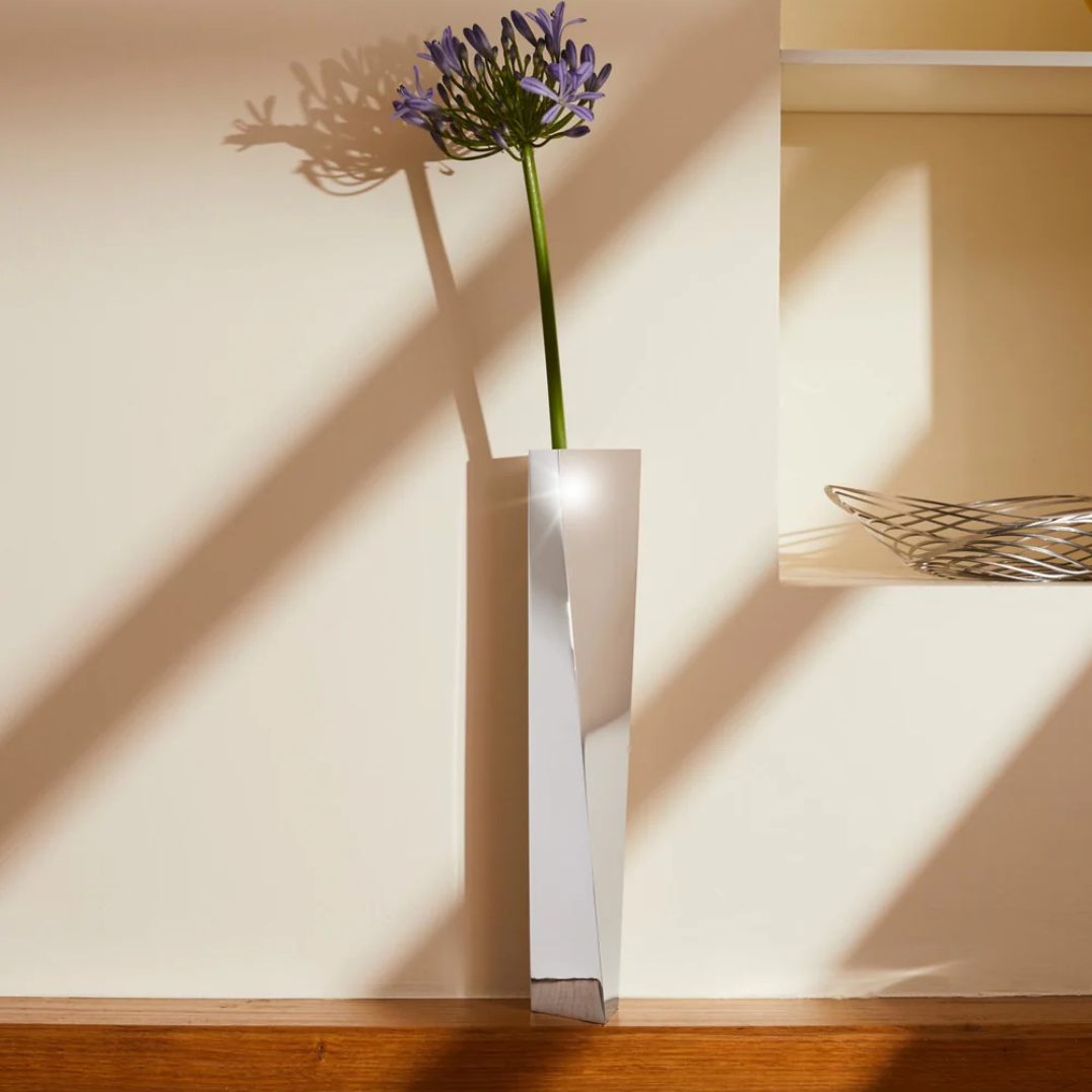 A tall silver vase with a single purple flower against a beige wall
