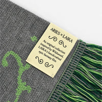 Close-up of a woven scarf with gray and green knit pattern and fringed edges, featuring a cream paper label that reads ARIES x LAIKA.