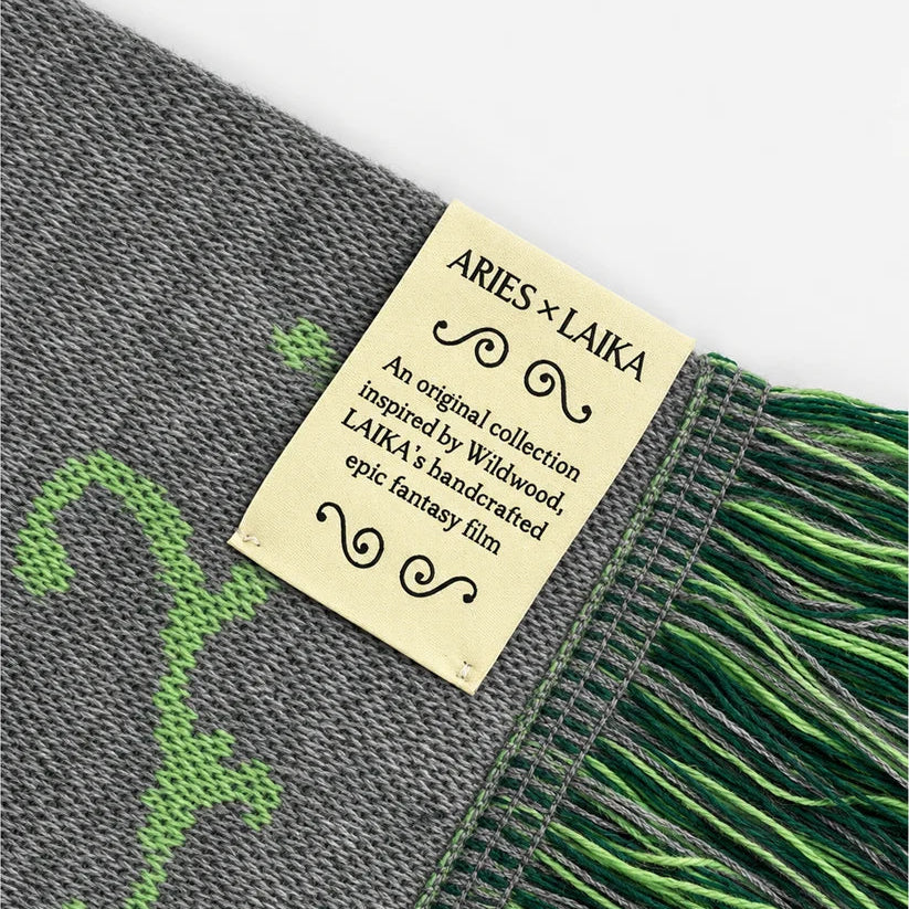 Close-up of a woven scarf with gray and green knit pattern and fringed edges, featuring a cream paper label that reads ARIES x LAIKA.