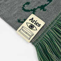 Close-up of a knitted scarf in gray and dark green with fringe, featuring a sewn-on cream label that reads 'Aries WILDMOOD'.