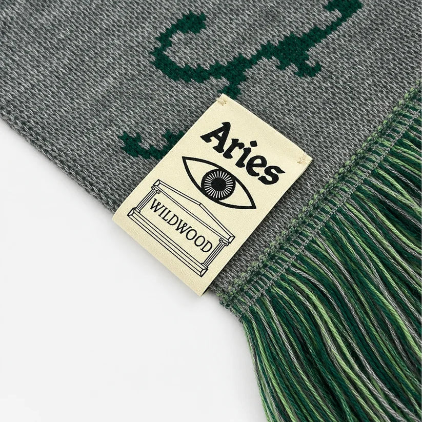 Close-up of a knitted scarf in gray and dark green with fringe, featuring a sewn-on cream label that reads 'Aries WILDMOOD'.