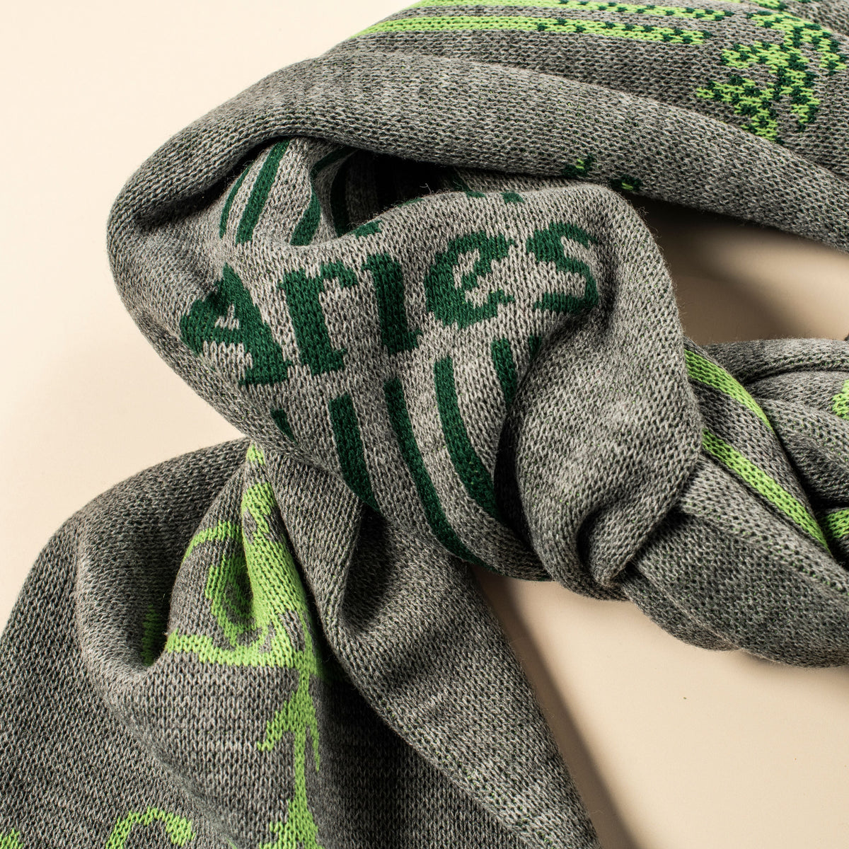 A close-up of a gray knit scarf with green woven lettering and pattern details, draped on a light beige background.
