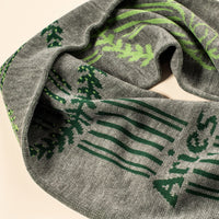 A close-up of a gray knit infinity scarf with a green patterned design featuring musical notes and abstract motifs, set against a plain light beige background.