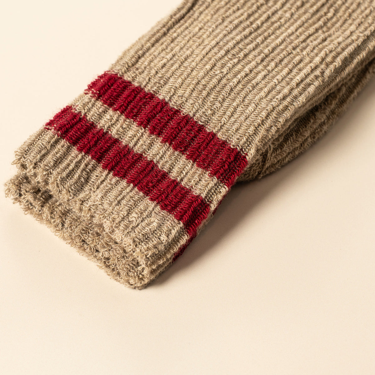 A close-up of a ribbed knit sock cuff in a heathered beige/tan color with two horizontal red stripes, photographed on a plain warm off-white background.
