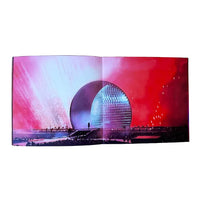 An open printed book or magazine spread photographed against a plain white background. The pages show a vivid architectural scene with a large metallic dome under dramatic pink, red, and blue lighting.