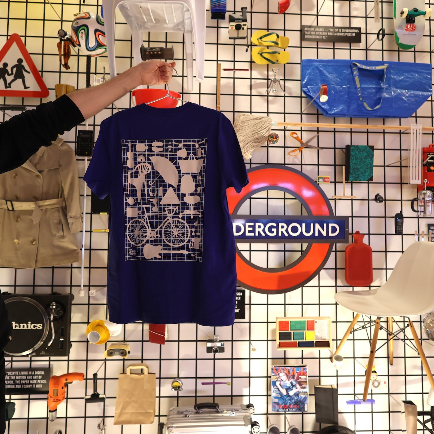 A picture of a blue t-shirt on a hanger held by a hand showing its back print in front of the Design Museum's Crowdsourced wall.