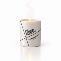 A white ceramic candle jar with a lit flame, featuring black text that reads 'the DESIGN MUSEUM' and 'CRANBOURN LONDON' on a clean white background.