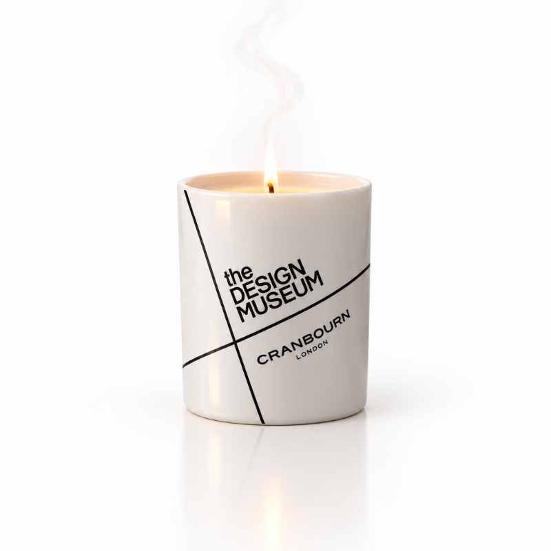 A white ceramic candle jar with a lit flame, featuring black text that reads 'the DESIGN MUSEUM' and 'CRANBOURN LONDON' on a clean white background.