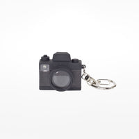 A black camera-shaped LED keychain with a realistic design, including a lens and flash, on a white background.