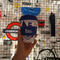 Hand holding a Design Museum cup in front of a wall with various objects