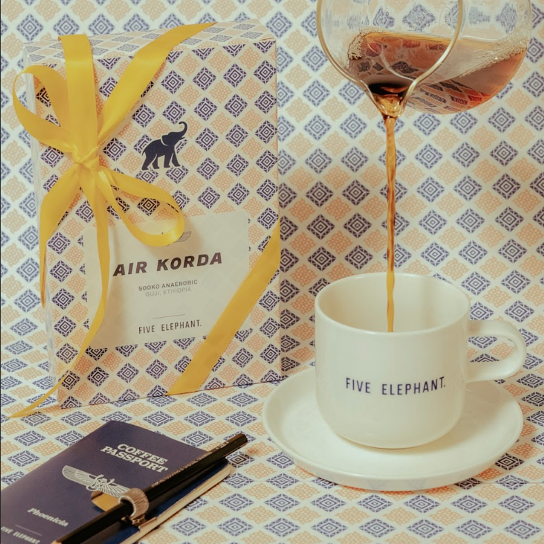 Hand pouring coffee into a 'Five Elephant' mug with a gift box and notebook in the background.