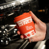 Hand holding a red coffee cup with 'The Design Museum' branding in front of a coffee machine.