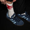 Person wearing blue Adidas sneakers with white stripes and a white sock with red stripes on a dark background