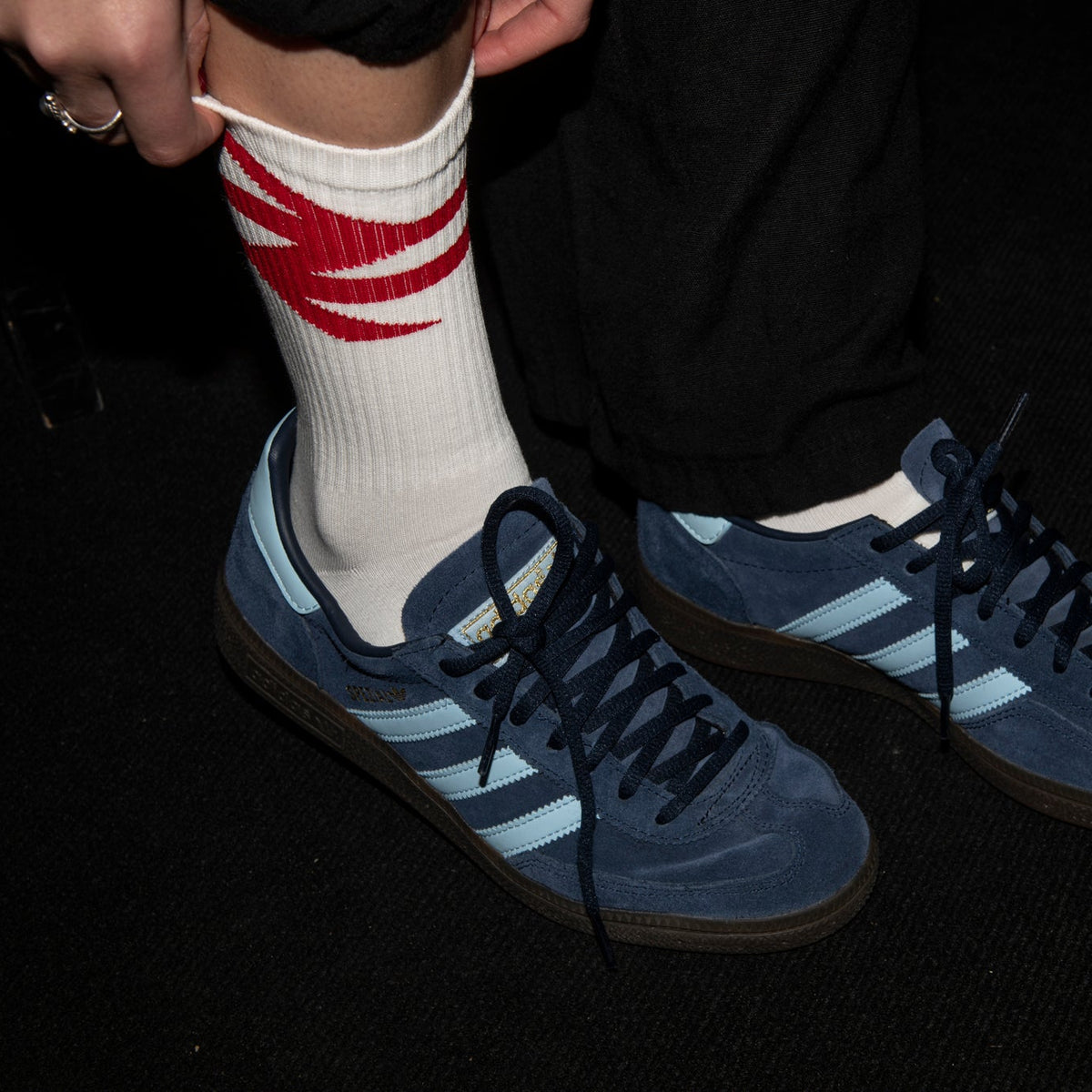 Person wearing blue Adidas sneakers with white stripes and a white sock with red stripes on a dark background