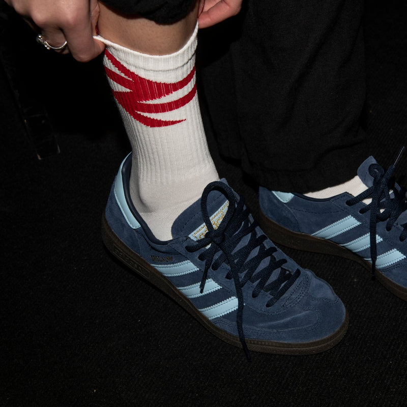 Person wearing blue Adidas sneakers with white stripes and a white sock with red stripes on a dark background.