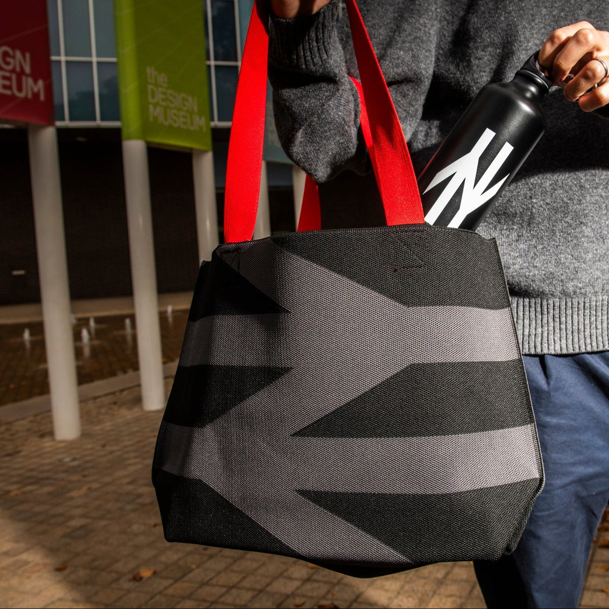 Person holding a black and gray geometric bag with red handles, standing in front of 'The Design Museum'.