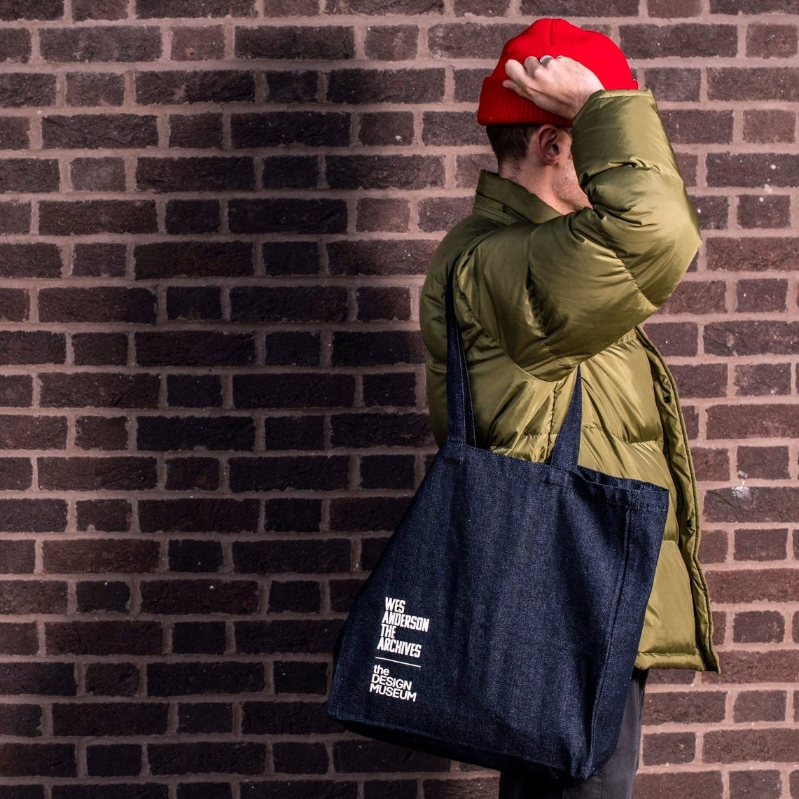 Person wearing a green jacket and red cap, holding a navy tote bag against a brick wall.