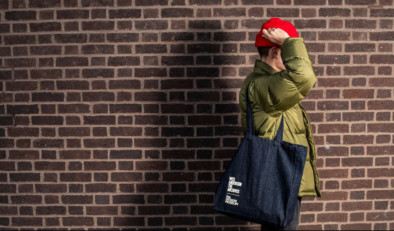 Person wearing a green coat and red hat against a brick wall, holding a navy tote bag.