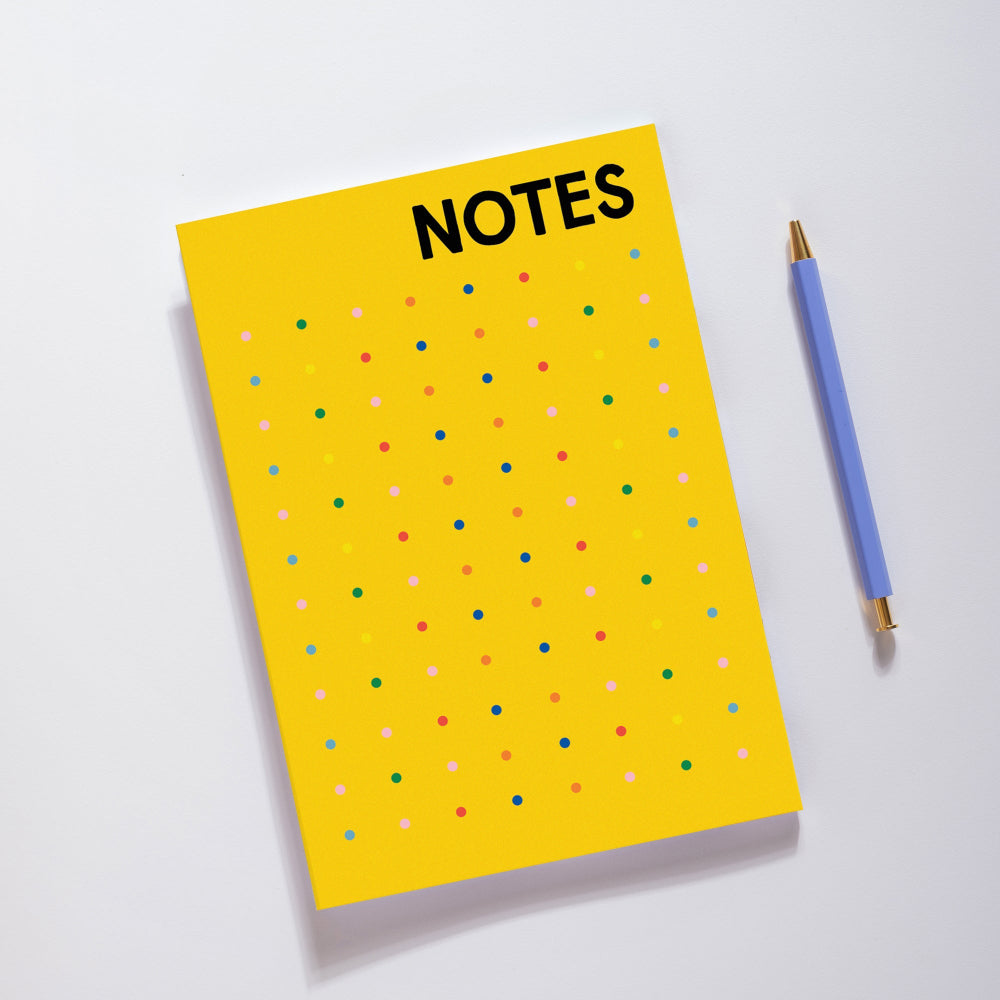 A bright yellow notepad with the word 'NOTES' in bold black letters at the top, decorated with a grid of small multicolored dots, shown on a white background next to a blue pen.