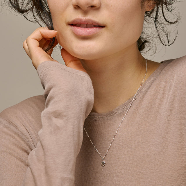A close-up of a woman wearing a delicate silver necklace with a small heart-shaped pendant, styled with a taupe long-sleeve top against a neutral beige background.
