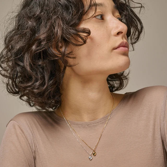 Woman with wavy hair wearing two heart necklaces against a neutral background