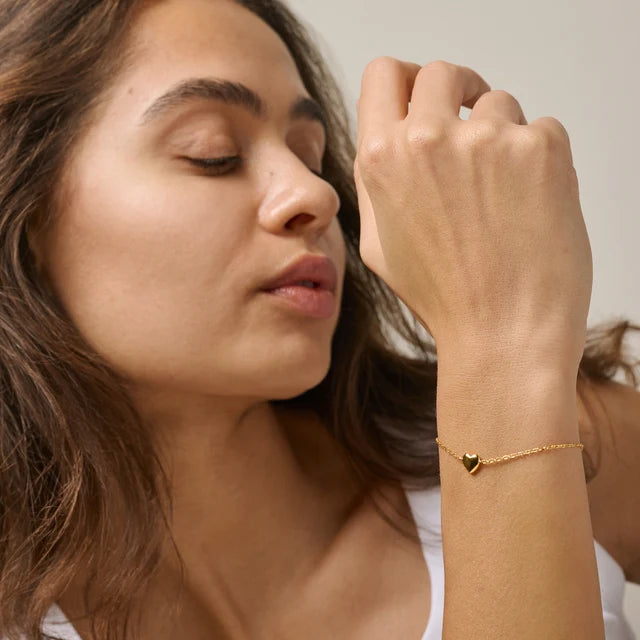 A close-up lifestyle shot of a woman wearing a delicate gold chain bracelet with a small heart-shaped charm on her wrist.