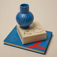 A styled jewelry display featuring gold and silver beaded necklaces, bracelets, and earrings arranged around a blue vase and two books. 