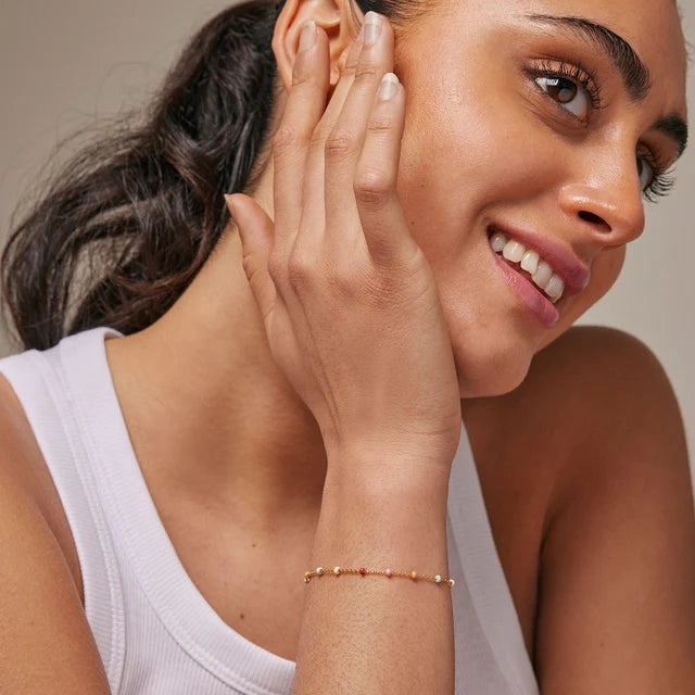 A close-up lifestyle shot of a woman wearing a delicate beaded bracelet on her wrist, smiling and touching her face against a soft beige background.