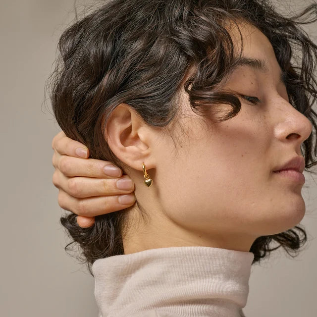 A close-up side profile of a woman with short dark curly hair wearing a small gold hoop earring with a gold heart charm, against a neutral beige background.