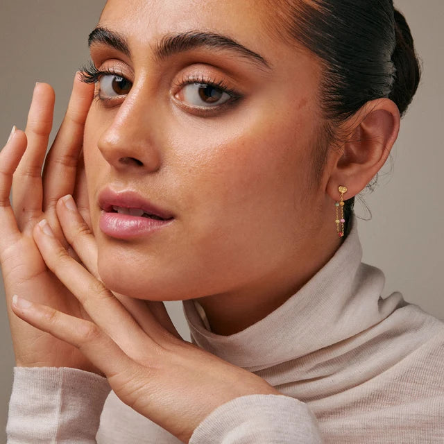 A close-up lifestyle shot of a woman wearing a delicate gold dangle earring with small stacked beads, posing with her hands near her face against a neutral beige background.